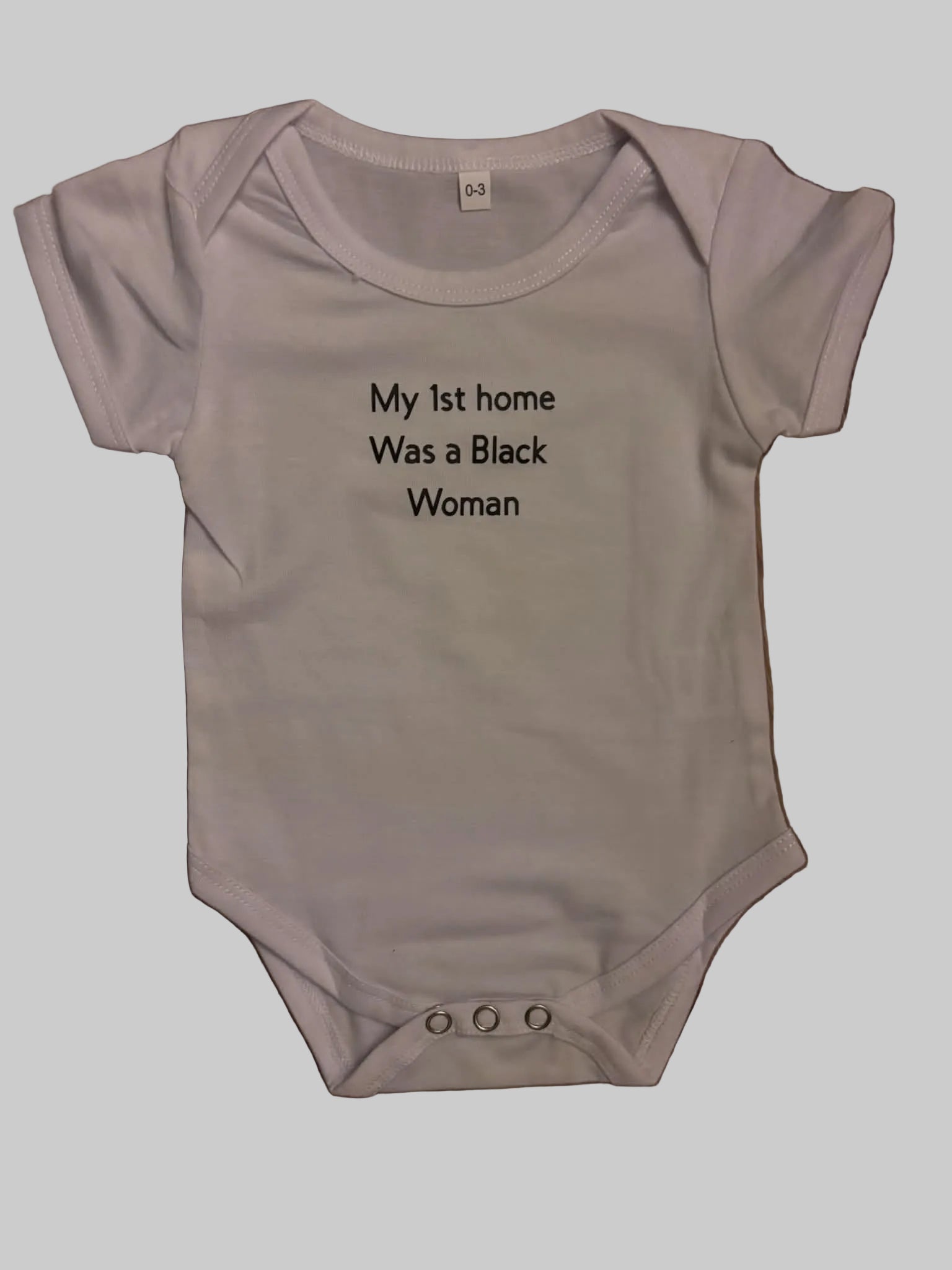 A white short-sleeve baby bodysuit with black printed text reading 'My 1st home Was a Black Woman' laid flat on a brown wooden floor.