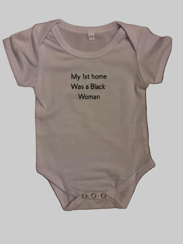 A white short-sleeve baby bodysuit with black printed text reading 'My 1st home Was a Black Woman' laid flat on a brown wooden floor.