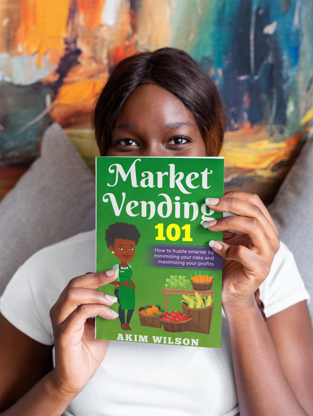 Person holding a book titled 'Market Vending 101' by Akim Wilson in front of a colorful painting.
